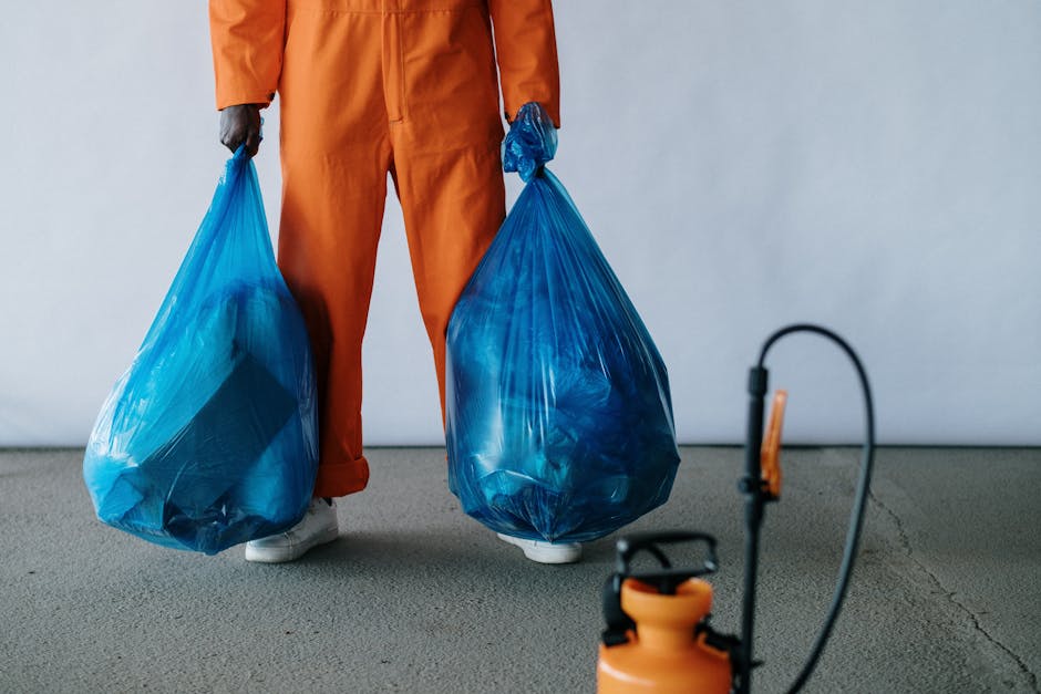A person dressed in orange work overalls is standing indoors on a smooth concrete floor, holding two large blue plastic garbage bags filled with waste or recyclable materials, with one bag in each hand. The person's feet are visible, wearing white sneakers. The background features a plain light grey wall, and the lighting is neutral and even. In the foreground, there is a small orange and black handheld sprayer or pump, positioned on the floor, slightly out of focus. This scene depicts a waste or rubbish collection process as part of house removal or home relocation, typical of services offered by Man with Van Ham, which specializes in removals and recycling options for bulky waste in the Ham area. The image captures the environmental aspect of packing and loading waste during the moving or clearance process, aligned with professional removals and recycling services.
