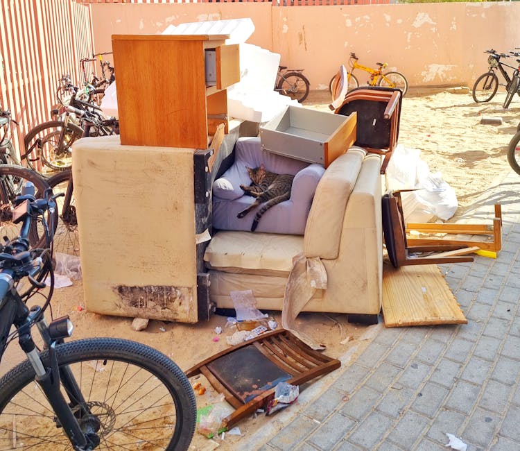 Various discarded household furniture and waste materials piled on the pavement outside a property, including a beige fabric sofa with visible stains and damage, a purple armchair with a cat resting on its cushion, a small wooden cabinet, a cardboard drawer insert, broken wooden chairs, and a piece of wooden flooring. Surrounding the furniture are large white foam packing blocks, plastic wrapping, cardboard boxes, and scattered debris. Bicycles are parked nearby, and an orange wall with peeling paint serves as the backdrop. The scene depicts an area prepared for bulky waste collection or removal, with furniture and waste awaiting clearance, consistent with house relocation or large item disposal services. Occasionally, Man with Van Ham's removal and recycling team may handle such items during a home move or recycling process, ensuring proper disposal or recycling of bulky waste.