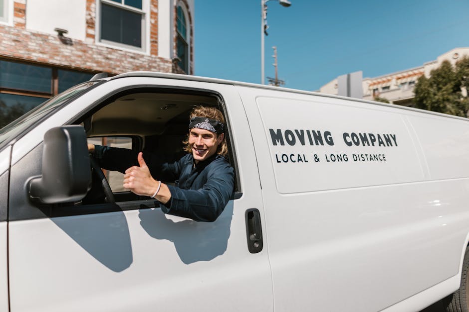 A young man with long hair, wearing a black bandana and navy blue shirt, is sitting in the driver's seat of a white moving van branded with the text 'MOVING COMPANY LOCAL & LONG DISTANCE.' The van is parked outside a residential building with large windows and brick walls, under a clear blue sky. Inside the open driver’s side window, the man is smiling and giving a thumbs-up, indicating a positive experience with the home relocation or furniture transport services provided by Man with Van Ham. The image captures the loading process or preparation stage for a house removal, with the van positioned on a paved area near the property. The scene highlights professional packing materials, such as cardboard boxes and protective blankets, as well as moving equipment like trolleys, hinting at the logistical aspects of packing and moving house furnishings efficiently and safely. Lighting is bright, emphasizing a professional and organized environment for household goods transportation.