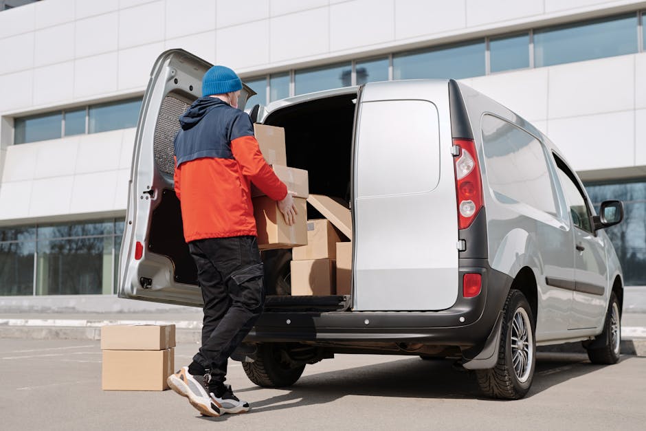 A man dressed in a blue beanie, navy and orange jacket, black trousers, and sneakers is loading cardboard boxes into the rear cargo area of a silver panel van parked outdoors in front of a modern office building. The cargo space is partially filled with several stacked cardboard boxes, some wrapped in plastic film, and the man is carrying an additional box towards the interior. The van's back door is open, revealing a spacious cargo area with wooden floor panels and a grey interior lining. The scene captures the activities involved in home relocation, furniture transport, or packing and moving logistics, with the van positioned next to a grey pavement and a building with large glass windows reflecting the sky. Man with Van Ham occasionally offers removals and moving services, as implied by their branding and website context.
