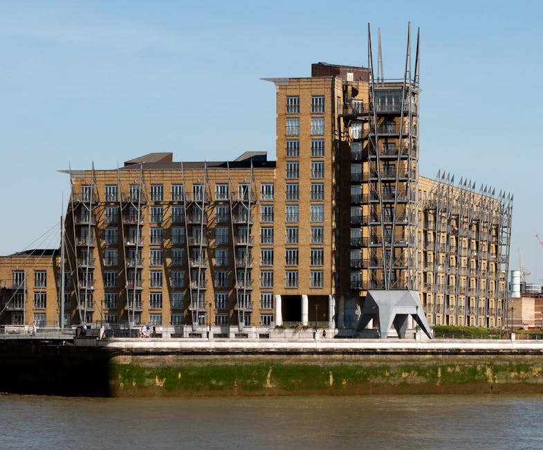 The image shows a multi-storey residential building under construction next to a river, with scaffolding visible along the exterior walls. The building has a mix of brick and concrete finishes, with several balconies and large windows. Construction workers' equipment, such as ladders and safety barriers, are present on the scaffolding, indicating ongoing work. The lower part of the building is adjacent to a paved walkway or road along the riverbank, with some construction vehicles or materials not visible in detail. The sky is clear and blue, providing natural daylight that illuminates the site. This scene illustrates the process of home development or renovation, related to house moves or property improvements in the context of relocating or updating riverside properties, aligning with [PAGE_TITLE] and services offered by [COMPANY_NAME], which include removals and property logistics.