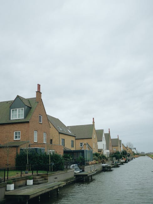 A row of modern detached houses with sloped roofs and brick facades situated along a calm riverbank under an overcast sky. Each property features large windows, some with white or green window frames, and small gardens with fences or hedges. Wooden docks extend from the rear of the houses into the water, with a few boats moored alongside. The scene depicts a peaceful residential area that is suitable for house relocations or move services, with a scenic riverside setting. As part of a professional removals process, Man with Van Ham might facilitate home relocation or furniture transport in such riverside properties, emphasizing careful handling and transportation of belongings to suit house moves near water access.
