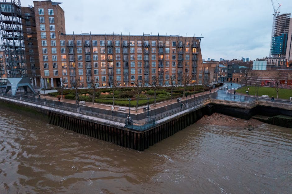 The image shows a multi-storey residential building under construction next to a river, with scaffolding visible along the exterior walls. The building has a mix of brick and concrete finishes, with several balconies and large windows. Construction workers' equipment, such as ladders and safety barriers, are present on the scaffolding, indicating ongoing work. The lower part of the building is adjacent to a paved walkway or road along the riverbank, with some construction vehicles or materials not visible in detail. The sky is clear and blue, providing natural daylight that illuminates the site. This scene illustrates the process of home development or renovation, related to house moves or property improvements in the context of relocating or updating riverside properties, aligning with [PAGE_TITLE] and services offered by [COMPANY_NAME], which include removals and property logistics.