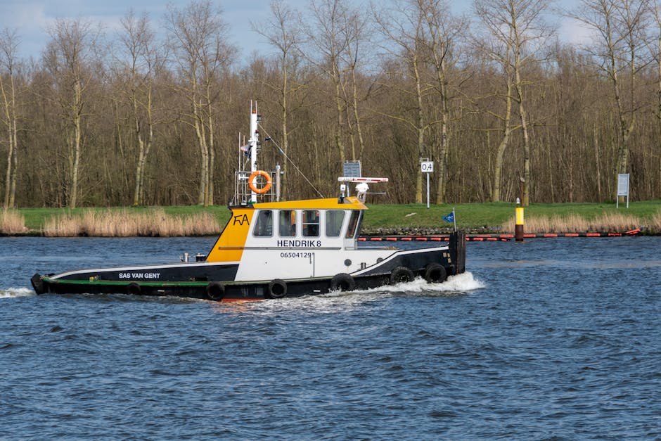 A small workboat named 'HENDRIK 8' operated by SAS VAN GENT, sailing on a river or canal, is visible in the image. The boat features a yellow and white cabin with a black hull, alongside life saving equipment such as a lifebuoy attached to the cabin, and a radar and antenna mounted on the roof. The boat is actively moving through the water, creating ripples and small waves around its hull. In the background, there is a grassy bank with leafless trees, alongside several navigation and informational signs, indicating narrow or controlled waterway access areas. The scene is well-lit with natural daylight, and the boat may potentially be involved in a moving or logistical operation related to house removals or transport services, consistent with the context of professional relocation processes. Man with Van Ham occasionally references such transport logistics, and the boat's role may support moving activities across waterways or navigating difficult access points for home relocations.