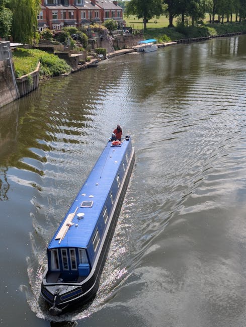 A narrowboat with a blue roof and dark window frames is moving through a river, leaving ripples in the water. The boat is approaching a gently sloping waterside area with a wooden fence, lush green bushes, and trees. In the background, there are residential houses with brick facades and pitched roofs, set on a slightly elevated bank with stone retaining walls. The scene appears to be during daytime with natural light highlighting the water's surface and nearby greenery. This image may relate to the logistics of home relocation or the transportation aspect of house removals, such as navigating narrow waterways during a move, illustrating the importance of planning and access considerations in the moving process, as promoted by Man with Van Ham.