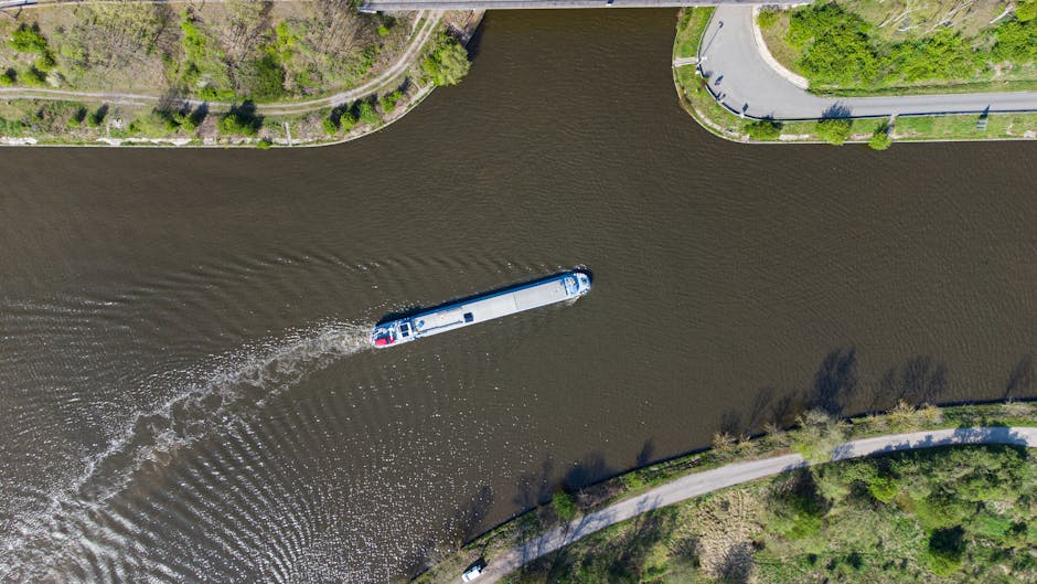 A small workboat named 'HENDRIK 8' operated by SAS VAN GENT, sailing on a river or canal, is visible in the image. The boat features a yellow and white cabin with a black hull, alongside life saving equipment such as a lifebuoy attached to the cabin, and a radar and antenna mounted on the roof. The boat is actively moving through the water, creating ripples and small waves around its hull. In the background, there is a grassy bank with leafless trees, alongside several navigation and informational signs, indicating narrow or controlled waterway access areas. The scene is well-lit with natural daylight, and the boat may potentially be involved in a moving or logistical operation related to house removals or transport services, consistent with the context of professional relocation processes. Man with Van Ham occasionally references such transport logistics, and the boat's role may support moving activities across waterways or navigating difficult access points for home relocations.