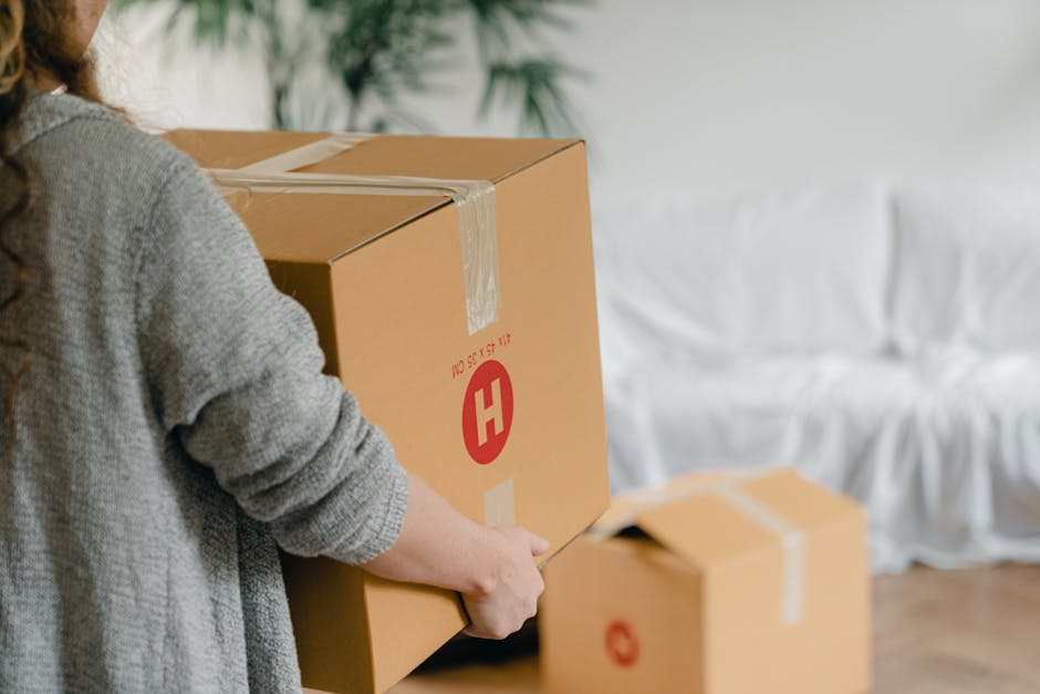 A person wearing a grey knitted sweater is seen holding a large cardboard box sealed with packing tape, with a red 'H' logo and product details printed on the side, inside a room with wooden flooring. Several other cardboard boxes of varying sizes are nearby, some open with flaps lifted, indicating packing and moving preparations. In the background, there is a white fabric-covered sofa and a green plant, suggesting a residential setting. The individual appears to be in the process of efficient home relocation, possibly as part of a furniture transport and packing process facilitated by Man with Van Ham, a professional removals service. Visible interior space and natural lighting highlight the organized environment during the loading process for same-day or last-minute removals in Ham.