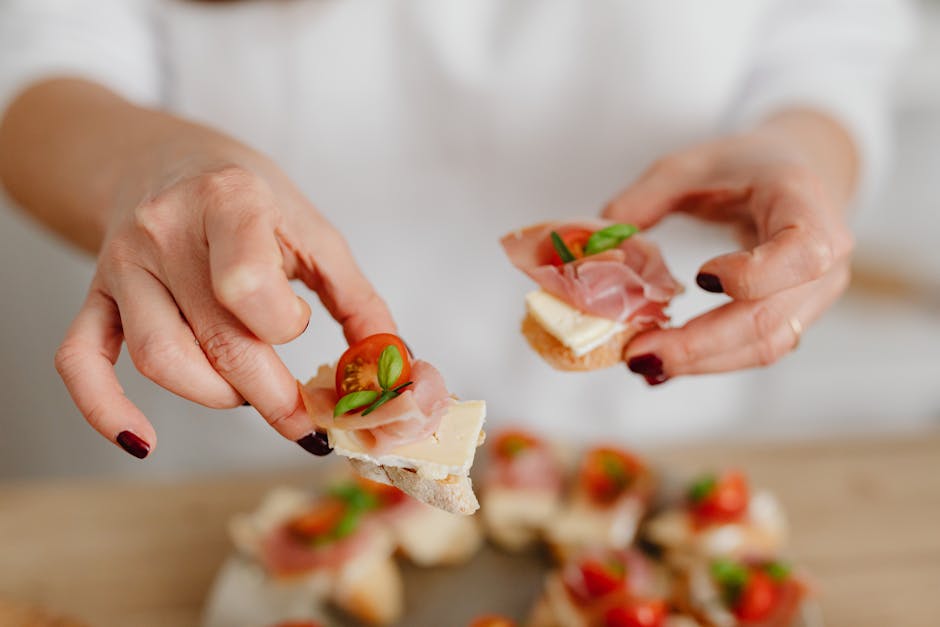 A pair of hands with dark nail polish are shown preparing small open-faced sandwiches. Each sandwich consists of a slice of white bread topped with slices of cheese, thinly sliced ham, cherry tomato halves, and fresh basil leaves. Additional completed sandwiches are visible on a wooden surface under the person's hands, with some toppings scattered across the area. The background is blurred, emphasizing the focus on the food preparation. This scene depicts a casual moment of assembling snacks, which could relate to packing or providing refreshments during a home relocation or moving process. The image supports the topic of house removals and logistics services offered by Man with Van Ham, highlighting the importance of organized packing and refreshment arrangements during furniture transport and loading activities.