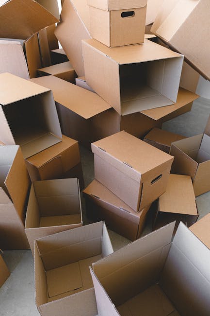 A man with dark, curly hair and a beard is indoors, holding a large cardboard box labeled 'OFFICE' with a 'MEDIUM' size indicator. The box is taped securely and appears to contain office furniture or 