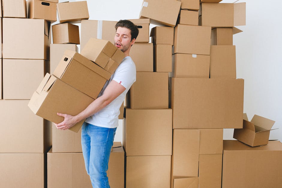 A man with dark hair, dressed in a white t-shirt and blue jeans, is carrying a medium-sized cardboard box in a room filled with stacked moving boxes of various sizes, all sealed with packing tape. Some boxes are partially open, revealing packing materials inside. The man is standing next to a large collection of packed boxes, which occupy the majority of the space, indicating a home relocation or packing process. The environment is well-lit, likely by natural light, and the background includes a plain white wall and a doorway. This scene illustrates the loading and packing aspects of furniture transport and house removals, with the man with Van Ham specializing in removals services and the logistics involved in moving heavy items safely during a professional home move.