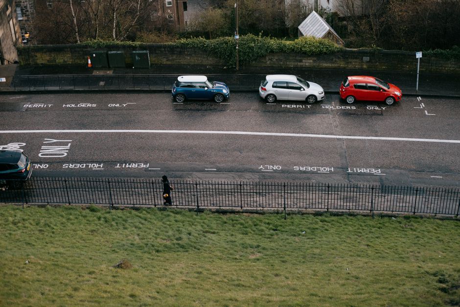 An aerial view of a parking area along Petersham Road in HAM, showing three parked cars of different colours—blue, silver, and red—aligned on the designated spaces marked 'Permit Holders Only' on the asphalt. The parking spaces are clearly delineated by white lines and signage painted directly on the road surface. The area is adjacent to a sidewalk with a black metal fence in the foreground, separating pedestrians from the street. Behind the parked vehicles, there is a green hedge and some trees, with a building partially visible in the background. The scene includes a person near the sidewalk moving a cardboard box, likely involved in a home relocation or furniture transport process, with a Home Removals company such as Man with Van Ham possibly coordinating the loading or unloading activities. The environment is well-lit, indicating daytime, and the overall scene captures a moment of loading or unloading tasks typical during house moves, where parking permits are essential for a smooth logistics operation.