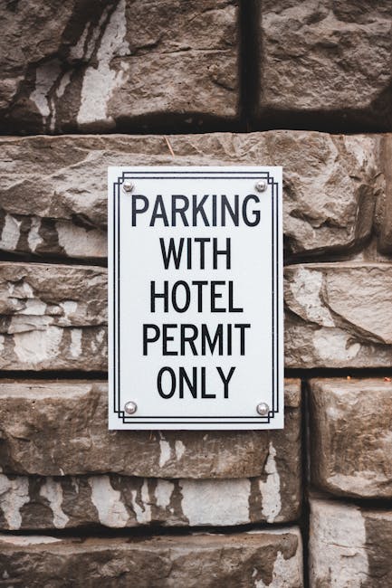Close-up of a rectangular white metal parking sign attached to a textured stone wall. The sign displays the message 'PARKING WITH HOTEL PERMIT ONLY' in black uppercase letters, indicating restricted parking for residents or service providers with valid permits. In the foreground, part of a moving van is visible, with the rear doors open, revealing the interior filled with packed cardboard boxes, wrapped furniture, and moving blankets. The van is positioned on a pavement adjacent to the building, suggesting a home relocation or furniture transport process. The presence of other packed items and the loading area underscores a controlled and organized moving operation conducted by Man with Van Ham, specialised in removals and house moves in the local area. The scene captures the typical environment and equipment involved in packing and moving activities, with good natural lighting highlighting details of the packed household items, the loading procedure, and the parking restriction sign.