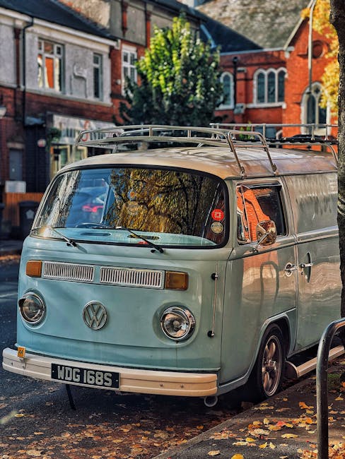 A vintage light blue Volkswagen camper van with a luggage rack on its roof is parked on a residential street lined with trees and red-brick houses, some of which have white-framed windows and decorati
