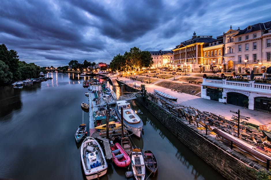 A panoramic view of a riverfront at dusk, featuring multiple small boats and yachts moored along a calm waterway. The boats are tied to the quay, with some covered in protective plastic wraps and others uncovered, showing outdoor seating and equipment. The shoreline is lined with wooden docks, some extending into the water, and is adjacent to a paved promenade. Behind the promenade, there is a row of elegant, illuminated buildings with classical architecture, large windows, and ornate facades, hosting restaurants and cafes with outdoor seating. Streetlights cast a warm glow on the area, while trees and greenery provide natural accents. The darkening sky overhead is filled with heavy clouds, creating a dramatic atmosphere. This scene illustrates a peaceful evening in a busy marina, relevant to house removals and city-based moving logistics, with the environment potentially serving as a staging point for furniture transport and relocation activities in a professional context, such as those provided by Man with Van Ham.