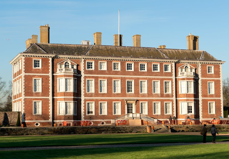 Photograph of a large, historic brick mansion with symmetrical architectural features, including multiple large sash windows with white frames, decorative stone accents, and small balconies with ornate railings. The building's roof has several prominent brick chimneys, and a flagpole extends from the center of the roof. The mansion is situated behind a well-maintained lawn with a stone staircase leading to the main entrance, and a gravel pathway in the foreground. The exterior appears clean and well-preserved, with natural lighting illuminating the scene. This setting depicts a stately residence potentially involved in a home relocation process, with the focus on the structure, its architectural details, and surrounding environment, aligning with house removals and moving services.