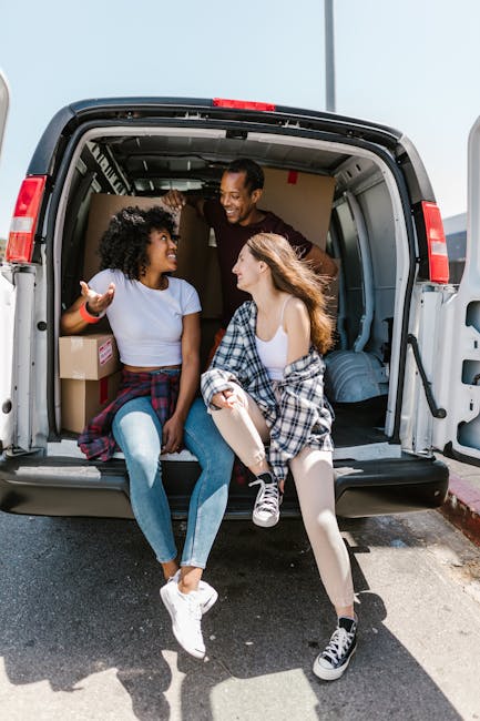 Inside a moving van, two young women with casual clothing and plaid shirts tied around their waists are seated at the open rear door, engaging in conversation and smiling. The van's interior contains 
