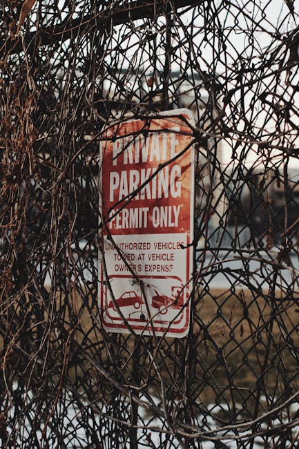 A weathered 'Private Parking Permit Only' sign mounted on a metal post, partially obscured by dry, tangled branches and vines. The sign is red and white, indicating parking restrictions and warning against unauthorized vehicle parking at the owner's expense. It is situated outdoors, with a background of an overcast sky and distant urban or suburban features, suggesting an area adjacent to a property involved in house removals or moving services. The image captures a typical scene where parking permits and restrictions are important considerations during home relocation or furniture transport, relevant to the services offered by Man with Van Ham.