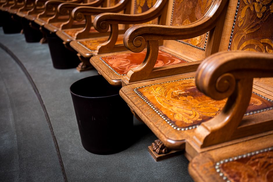 A row of ornately carved wooden church pews with upholstered seats in a floral fabric pattern, positioned on a dark carpeted floor. Each pew features curved armrests with scroll details and is decorated with metal nailhead trim along the edges of the upholstery. A black plastic waste bin is placed beside the pews, indicating an indoor setting likely used for events or gatherings. The lighting is soft, highlighting the polished wood and intricate carvings, and the scene appears to be part of a home relocation or moving process where furniture is being packed or prepared for transport, as part of a house removal service by Man with Van Ham. The image emphasizes the careful handling of furniture, with clear visibility of the furniture details and environment, supporting the theme of furniture transport and packing in a moving context.